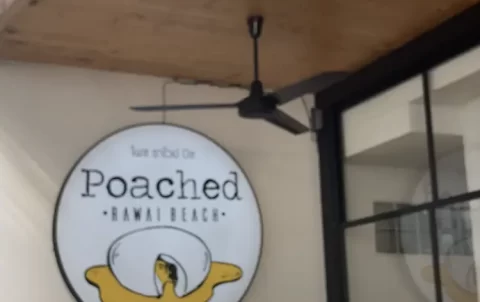 A hand holding a large plate of pulled pork eggs benedict with pomegranate seeds and mint on top, in front of the sign for Poached Breakfast Cafe, one of the best breakfast spots in Rawai, Phuket, Thailand