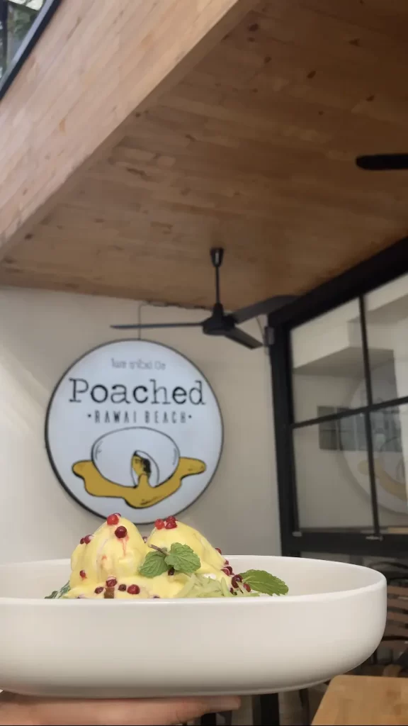 A hand holding a large plate of pulled pork eggs benedict with pomegranate seeds and mint on top, in front of the sign for Poached Breakfast Cafe, one of the best breakfast spots in Rawai, Phuket, Thailand