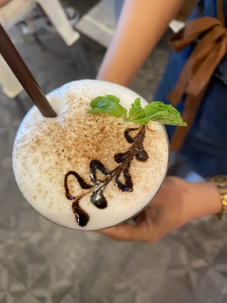 An overhead shot of an iced cappucino with a mint leaf on top, held by a server at the best coffeehouse in Rawai: Poached Breakfast Cafe, Rawai, Phuket, Thailand