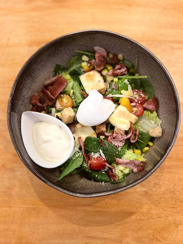 Close up of a Caesar salad topped with bacon, croutons, a poached egg and a side of Caesar dressing. A delicious option at one of the best places to eat in Rawai: Poached Breakfast Cafe, Rawai, Phuket, Thailand