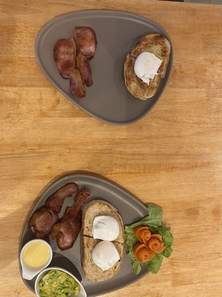 A plate with one piece of sourdough, a poached egg and 2 slices of bacon next to another plate with bacon, poached eggs, smoked salmon, smashed avocado, hollandaise sauce and sourdough bread at Poached Breakfast Cafe Rawai, Phuket, Thailand. The best iced coffee in Rawai is served here - and the best food too!