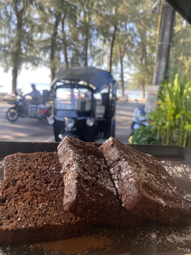 Warm banana bread in the foreground, sprinkled with cinnamon and icing sugar with a view of a traditional Thai tuk tuk and the beach in the background. One of the best backpacking activities in Rawai is right here at Poached Breakfast Cafe Rawai, Phuket, Thailand
