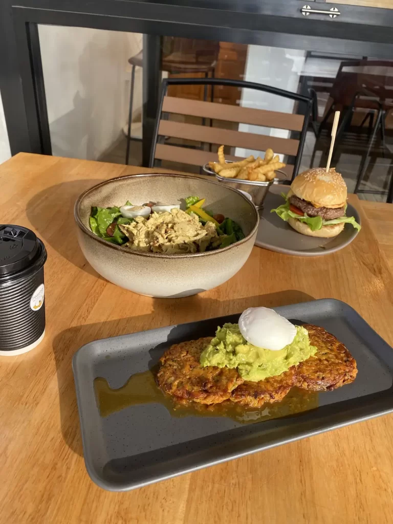 Burger and fries in the background next to a large bowl of curried chicken salad, a coffee in a takeaway cup and a plate of zucchini fritters topped with sweet chilli sauce, smashed avocado and a poached egg at one of the best tourist attractions in Naiharn at Poached Breakfast Cafe Rawai, Phuket, Thailand