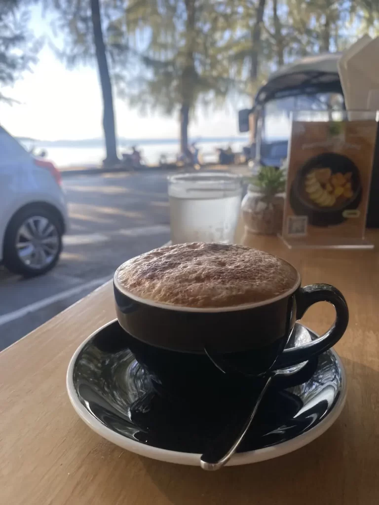 Frothy cappuccino on a wooden table in the foreground. There is a glass of cold water in the background and a view of the ocean with islands in the distance. One of the best backpacking activities in Rawai is right here at Poached Breakfast Cafe Rawai, Phuket, Thailand