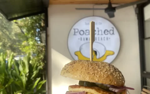 The image is focused on a halloumi bagel topped with lettuce, tomato, and onion with a side salad. In the background is a ceiling fan and the Poached Breakfast Cafe sign. One of the best backpacking activities is right here at Poached Breakfast Cafe Rawai, Phuket, Thailand