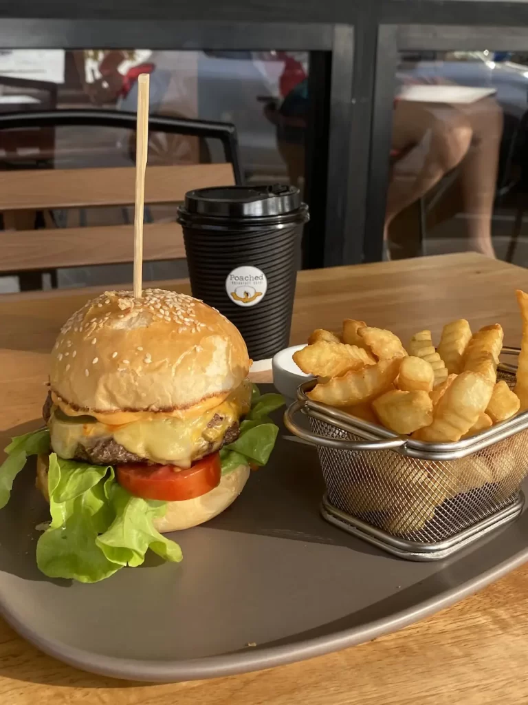 Juicy burger with 2 types of melty cheese, jalapenos, lettuce and tomato next to a basket of crispy crinkle fries and a take away coffee. Only at Poached Breakfast Cafe Rawai, Phuket, Thailand - the best Western breakfast in Rawai!