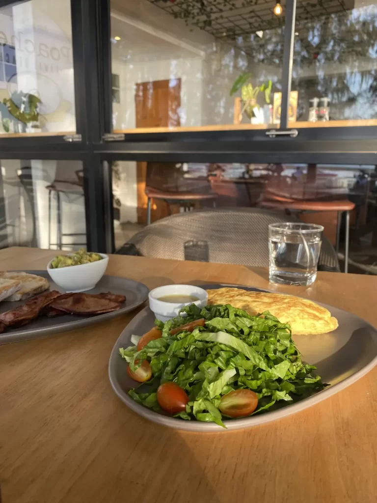 Salad on a plate with an omelette and salad dressing. There is a plate on the side with bacon, sourdough bread and smashed avocado. All this at Poached Breakfast Cafe Rawai, Phuket, Thailand. The perfect meal to fuel you up before you hit up one of the Muay Thai gyms in Rawai.