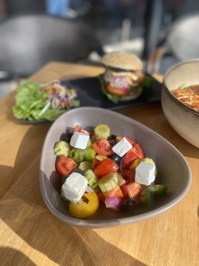 Greek salad in the foreground and a burger in the background on a table in the sunshine. All this at the best place to start when you're wanting to explore the walking routes in Rawai: Poached Breakfast Cafe Rawai, Phuket, Thailand