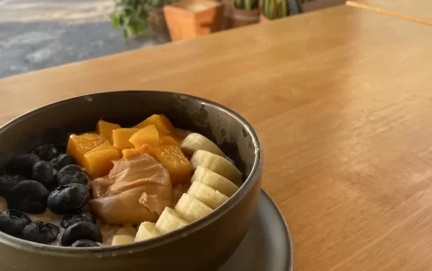 A bowl of vegan porridge on a table outdoors at Poached Breakfast Cafe Rawai, Phuket, Thailand. The porridge is topped with blueberries, mango, banana and peanut butter. The perfect meal to fuel you up before you hit up one of the Muay Thai gyms in Rawai.