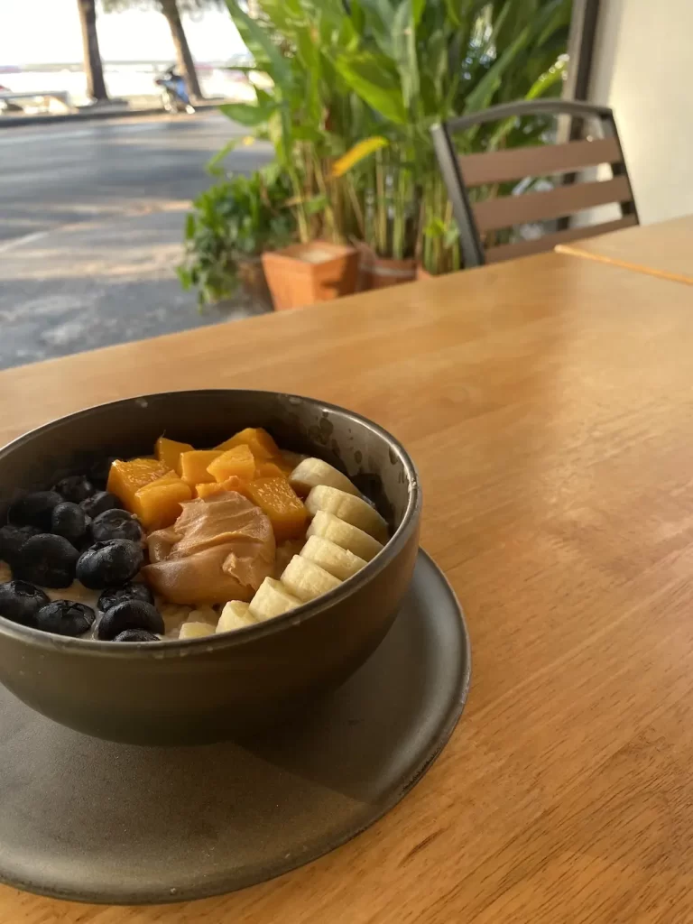 A bowl of vegan porridge on a table outdoors at Poached Breakfast Cafe Rawai, Phuket, Thailand. The porridge is topped with blueberries, mango, banana and peanut butter. The perfect meal to fuel you up before you hit up one of the Muay Thai gyms in Rawai.