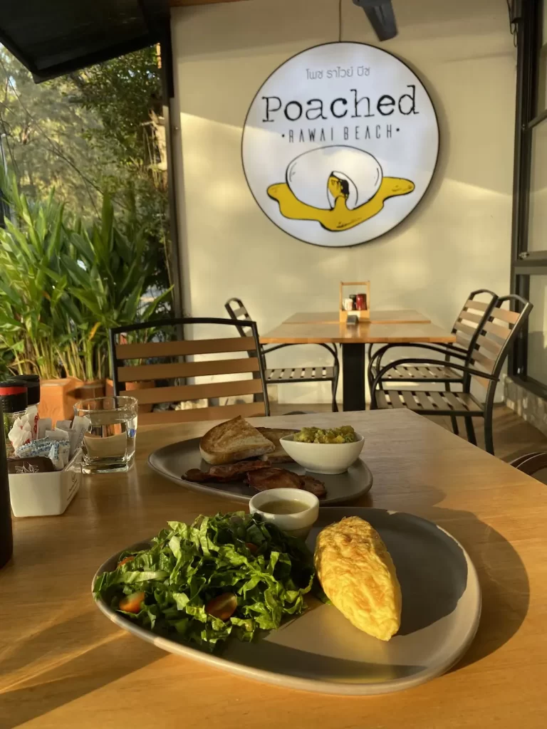 Healthy omelette with salad and dressing in the foreground, with sourdough bread, bacon and smashed avocado behind it. There is another table and a large outdoor sign in the background. All this at the spot with the best ocean view in Rawai: Poached Breakfast Cafe, Rawai, Phuket, Thailand