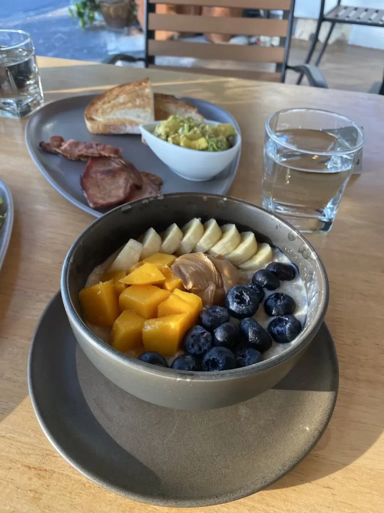 Bowl of protein porridge with mango, banana, peanut butter and blueberries and a side of sourdough bread, bacon and smashed avo. Only at the best spot for an ocean view in Rawai: at Poached Breakfast Cafe, Rawai, Phuket, Thailand