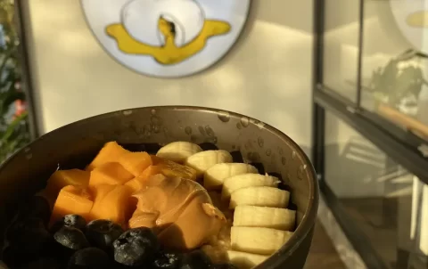 A hand holding up a warm vegan porridge bowl topped with peanut butter, banana, mangoes and berries at the place with the best cafe culture in Nai Harn: Poached Breakfast Cafe Rawai, Phuket, Thailand