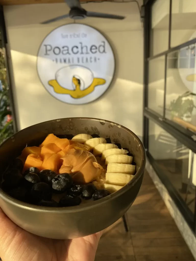 A hand holding up a warm vegan porridge bowl topped with peanut butter, banana, mangoes and berries at the place with the best cafe culture in Nai Harn: Poached Breakfast Cafe Rawai, Phuket, Thailand