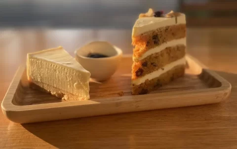 Cheesecake on the left, a small pot of blueberry jam in the middle and a layered carrot cake, all on a wooden plate. The plate is on a sunlit table opposite the beach in our beautiful outdoor seating in Rawai at Poached Breakfast Cafe Rawai, Phuket, Thailand