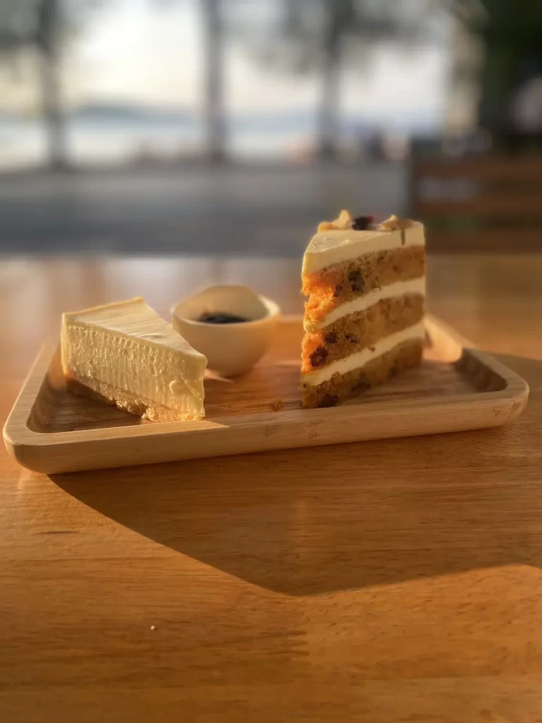 Cheesecake on the left, a small pot of blueberry jam in the middle and a layered carrot cake, all on a wooden plate. The plate is on a sunlit table opposite the beach in our beautiful outdoor seating in Rawai at Poached Breakfast Cafe Rawai, Phuket, Thailand