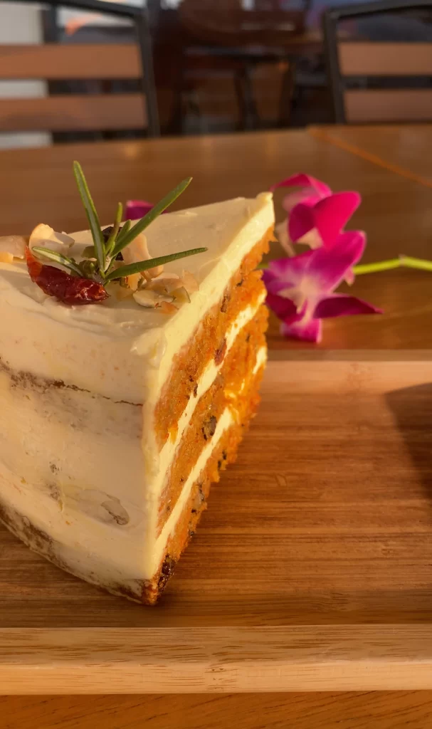 A piece of carrot cake on a table outdoors with a orchid in the background at the cafe with the best tea in Rawai: Poached Breakfast Cafe Rawai, Phuket, Thailand