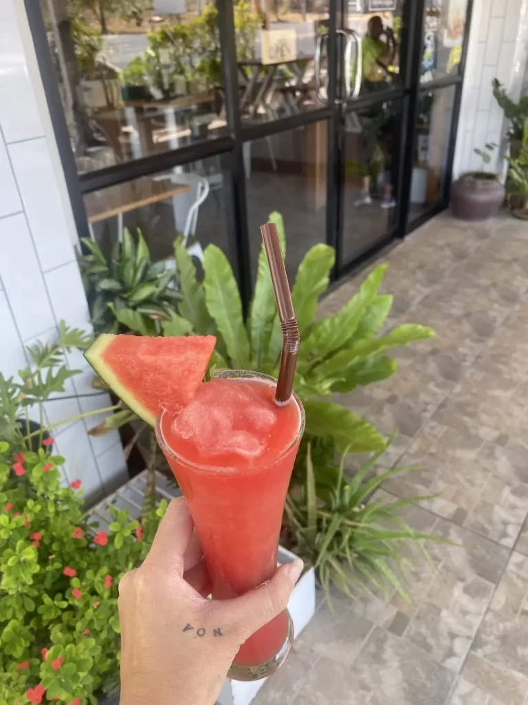 Someone holding the best watermelon smoothie near Rawai with foliage and glass doors in the background. Only at Poached Breakfast Cafe, Rawai, Phuket, Thailand