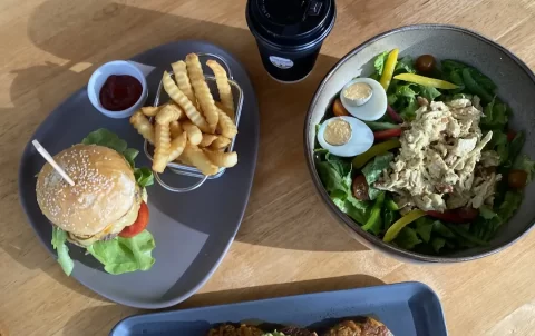 Curried chicken salad in a bowl with a boiled egg, a coffee in a takeaway cup, a plate of fritters with smashed avocado and a poached egg and a burger with fries and ketchup. Only at the spot with the best caesar salad near Nai Harn, Poached Breakfast Cafe Rawai, Phuket, Thailand