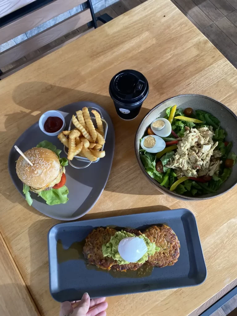 Curried chicken salad in a bowl with a boiled egg, a coffee in a takeaway cup, a plate of fritters with smashed avocado and a poached egg and a burger with fries and ketchup. Only at the spot with the best caesar salad near Nai Harn, Poached Breakfast Cafe Rawai, Phuket, Thailand