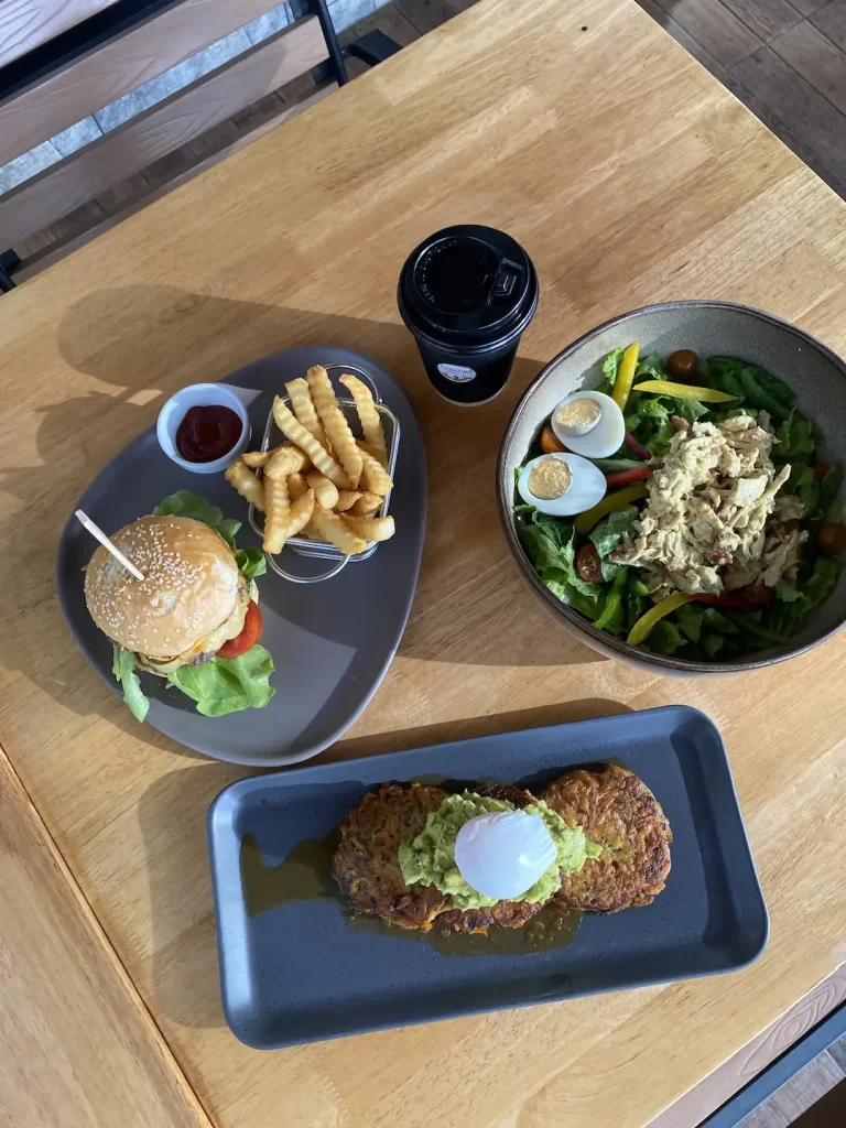 Burger with fries and ketchup, a coffee in a takeaway up, a curried chicken salad and sweet potato fritters with a poached egg at one of the most instagrammable spots near nai harn: Poached Breakfast Cafe Rawai, Phuket, Thailand
