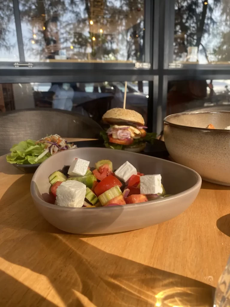 Greek salad in the foreground and a burger in the background. Only at the spot with the best caesar salad near Nai Harn, Poached Breakfast Cafe Rawai, Phuket, Thailand