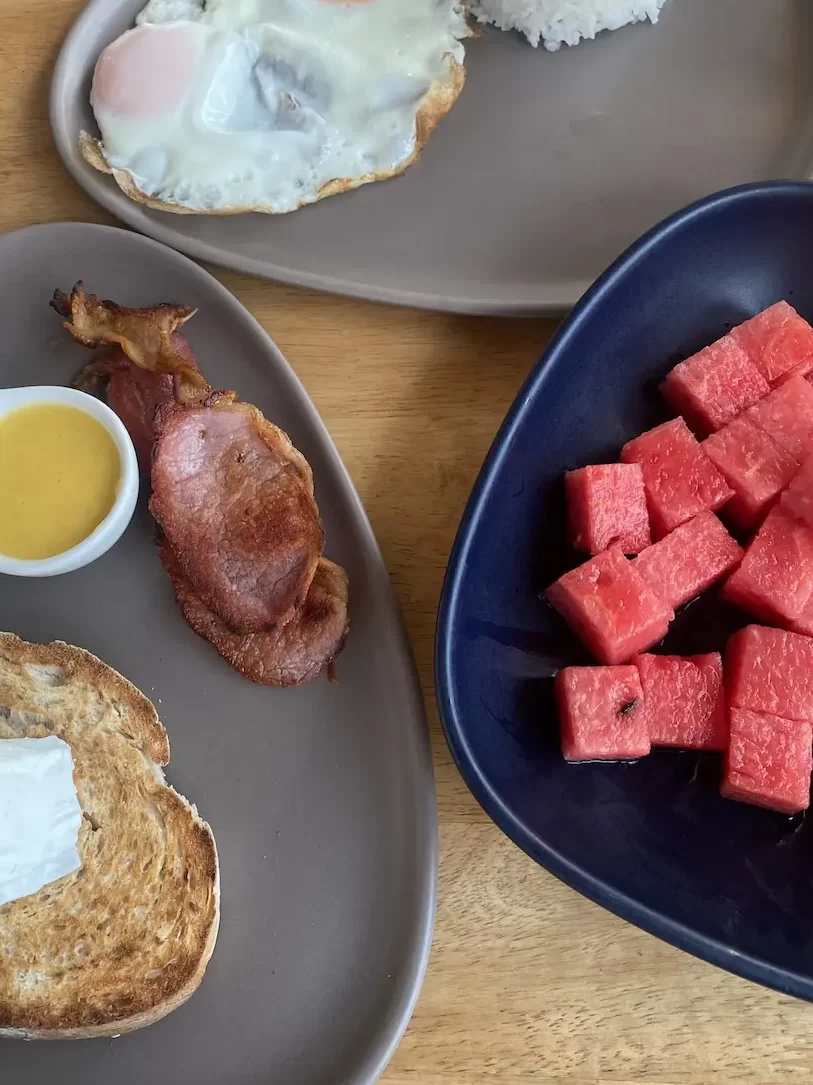 3 plates. One has 2 fried eggs, another has watermelon and another has 2 slices of sourdough, a bowl of smashed avocado, fried bacon, 2 poached eggs and hollandaise sauce. All at the best place to eat when you're staying at one of the hostels in Rawai: Poached Breakfast Cafe Rawai, Phuket, Thailand